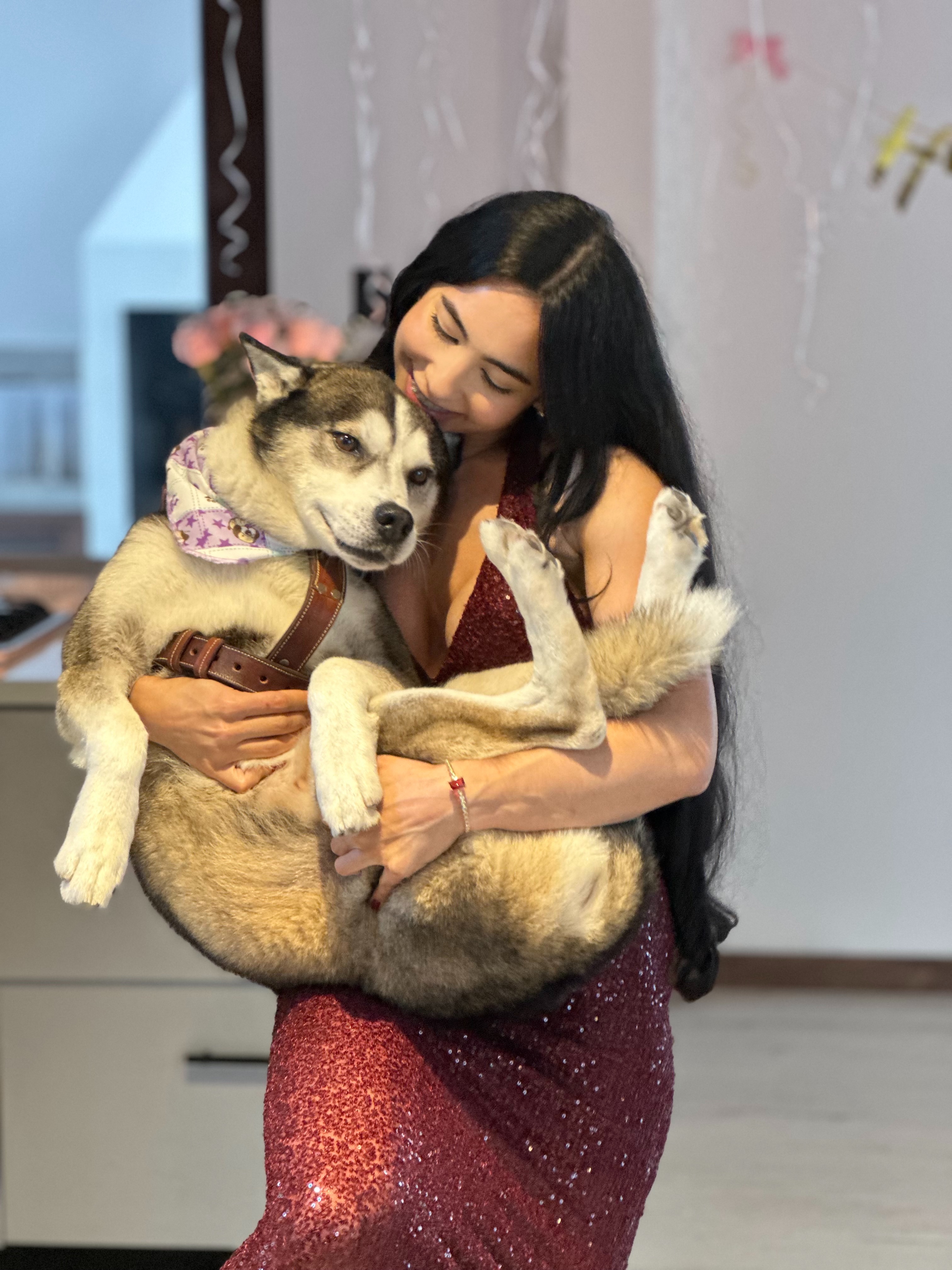 Maritza in a sparkly red dress embracing Nash the Siberian Husky, who wears a purple bandana, indoors.