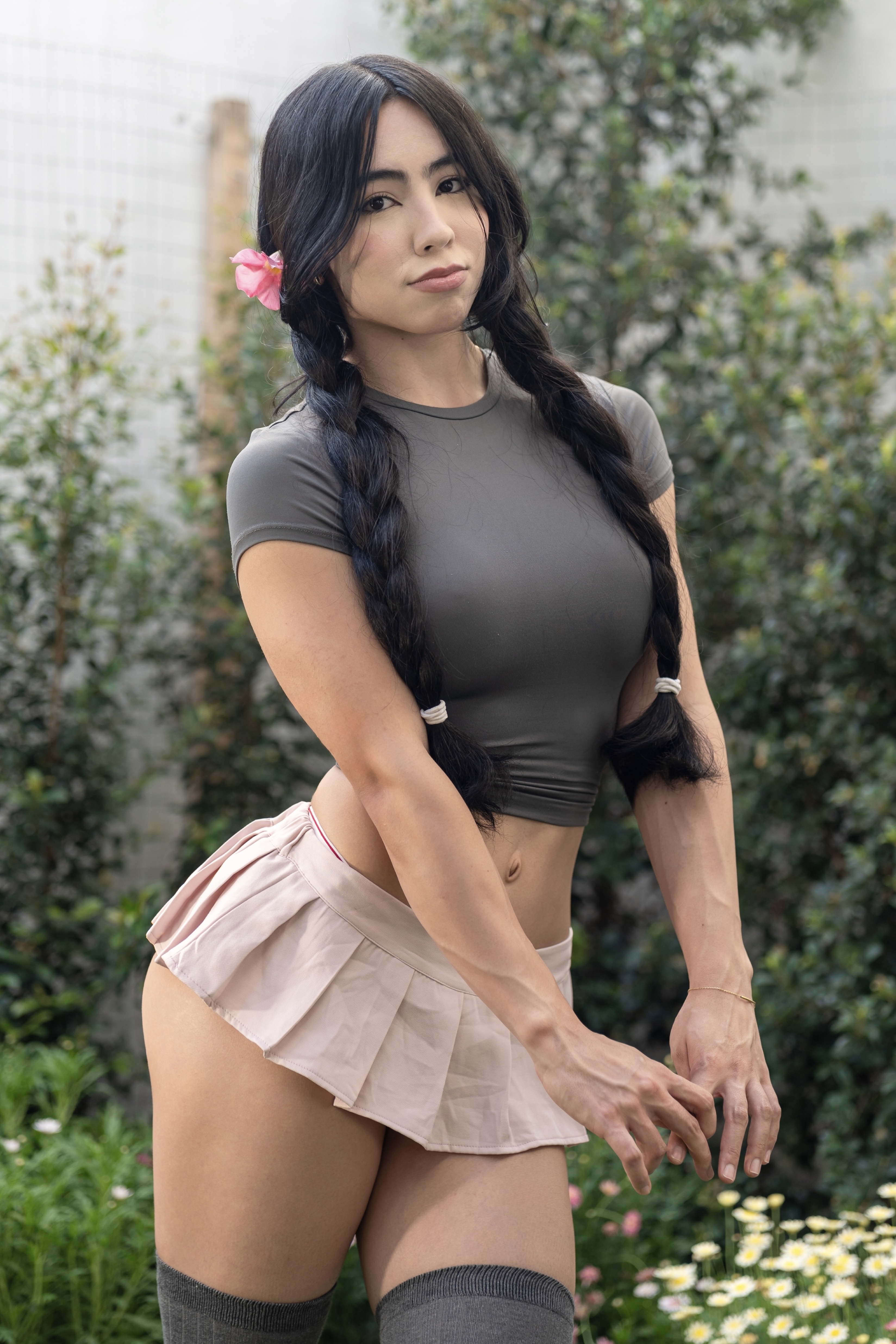 Maritza with braided hair and a pink flower, wearing a crop top and skirt, standing outdoors among green foliage.